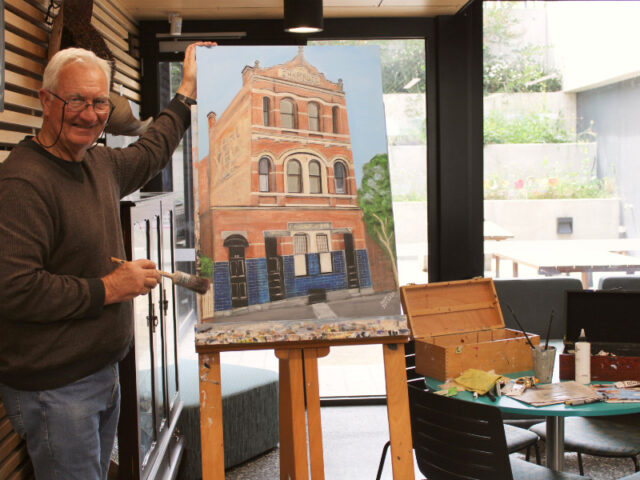 man with brush standing next to painting of old building on easel with paint supplies
