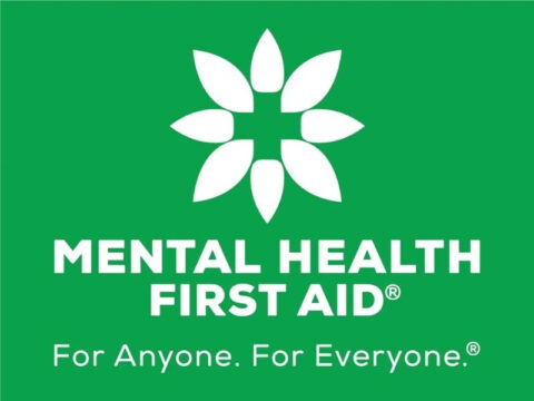 A Mental Health First Aid® logo on a green background. The logo features a white geometric symbol resembling a flower or star, composed of eight oval-shaped petals arranged in a circular pattern. At the center of the design, the negative space forms a white cross, symbolizing aid and support. Below the symbol, bold white capital letters spell out "MENTAL HEALTH FIRST AID®." The tagline "For Anyone. For Everyone.®" is written beneath in white sentence case.
