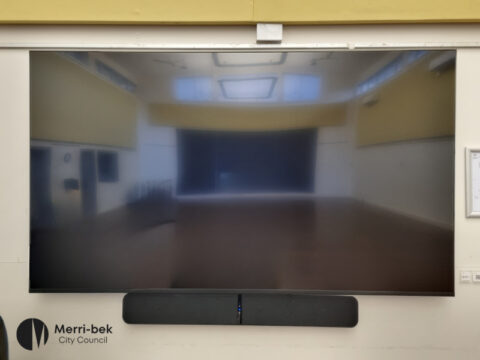 A large 100-inch TV mounted on a wall in a hall, reflecting the spacious room in front of it. A black soundbar is installed below the screen. The hall has high ceilings, windows, and light yellow walls. Merri-bek City Council logo is featured in the bottom left corner.