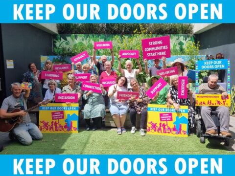 Group photo of community members holding signs with words like 'connection,' 'friendship,' and 'inclusion' in support of the 'Keep Our Doors Open' campaign.